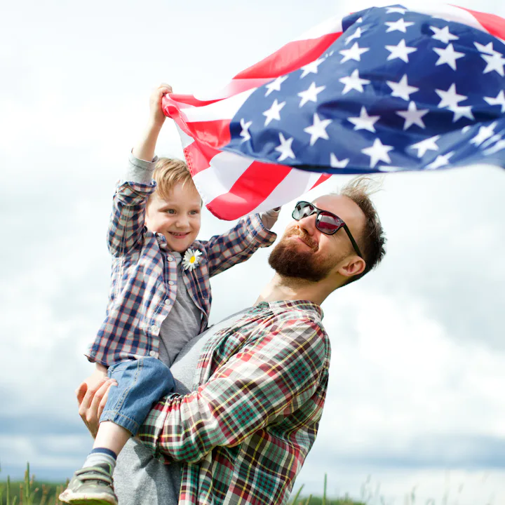 A man holding a young child who waves an American flag in a grassy field under a cloudy sky.
