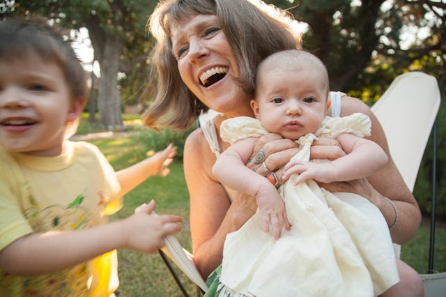 Grandmother laughing outdoors with a baby in her lap and a young boy in motion on her side.