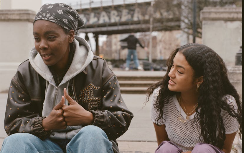 Two young women sitting on a step, one speaking animatedly to the other, in a casual, urban setting.