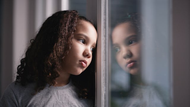 Young girl with curly hair looking thoughtfully out of a window, her reflection visible in the glass...