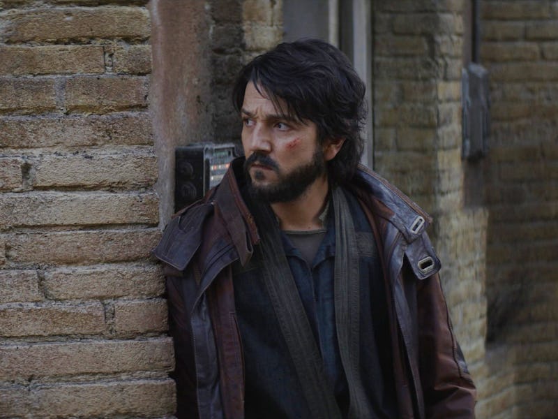 A rugged man with a beard stands against a textured brick wall, looking intently off to the side, su...