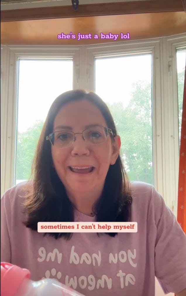 A woman with brown hair and glasses smiles at the camera, wearing a light purple shirt with humorous text. Bright windows are visible in the background.