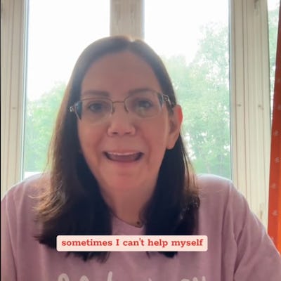 A woman with brown hair and glasses smiles at the camera, wearing a light purple shirt with humorous text. Bright windows are visible in the background.