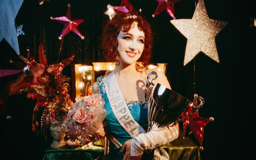 A joyful contestant in an elegant blue gown holds a trophy and bouquet, standing against a backdrop ...