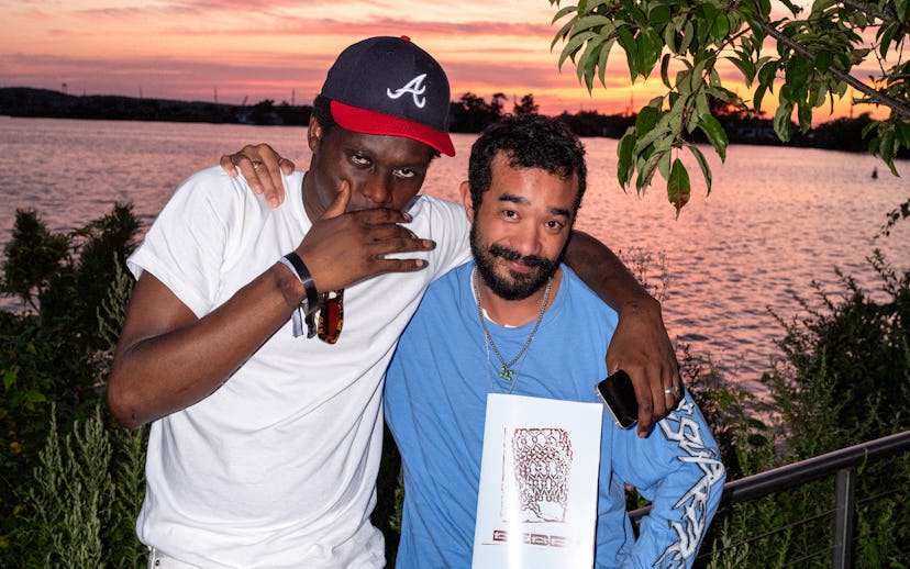 Two men pose for a photo by a waterfront at sunset, one holding a printed artwork. The background fe...