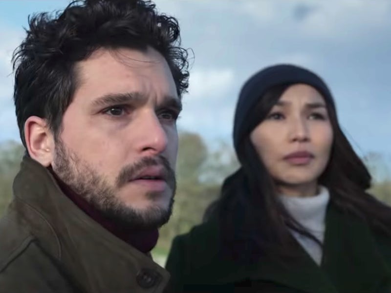 A man and woman stand outdoors, looking into the distance. The man has curly hair and is wearing a j...