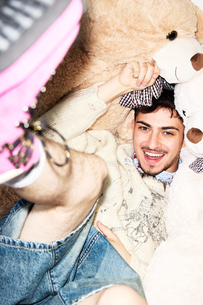 A young man smiles widely while surrounded by large teddy bears. He wears a cozy sweater and denim s...