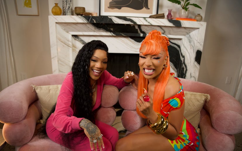 Two women are seated on a pink velvet sofa, smiling and laughing. One has long black hair and a pink...