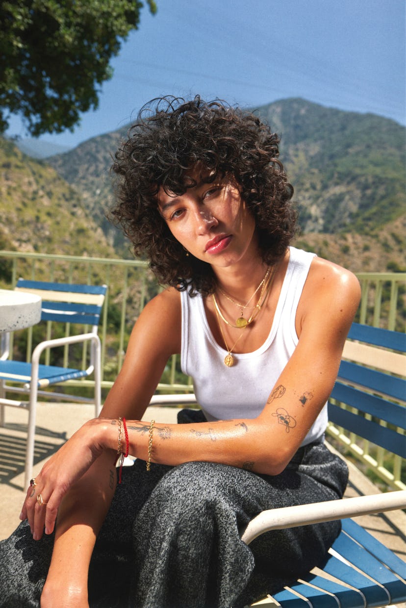 A young woman with curly hair sits on a blue and white chair, wearing a tank top and patterned pants...