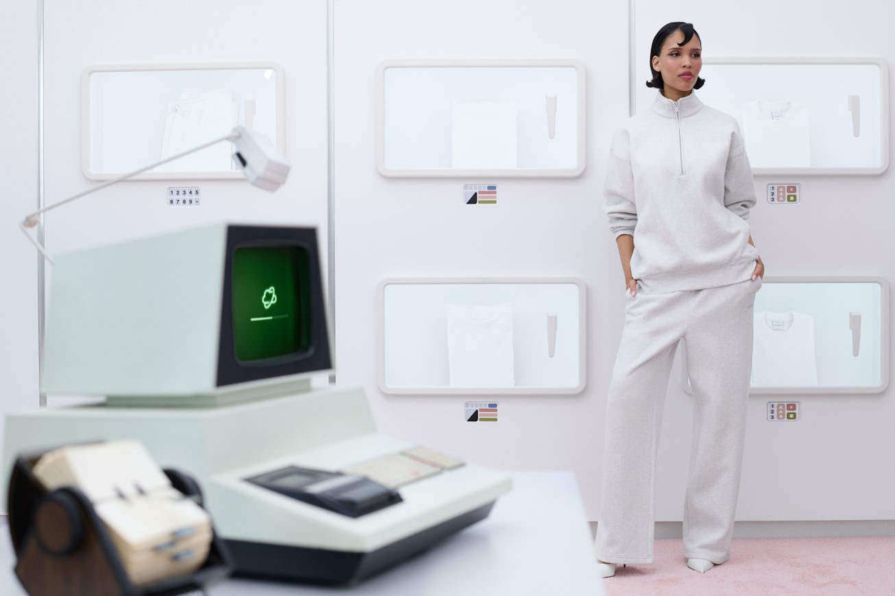 A stylish model stands beside a vintage computer in a modern exhibit. White walls display minimalist...