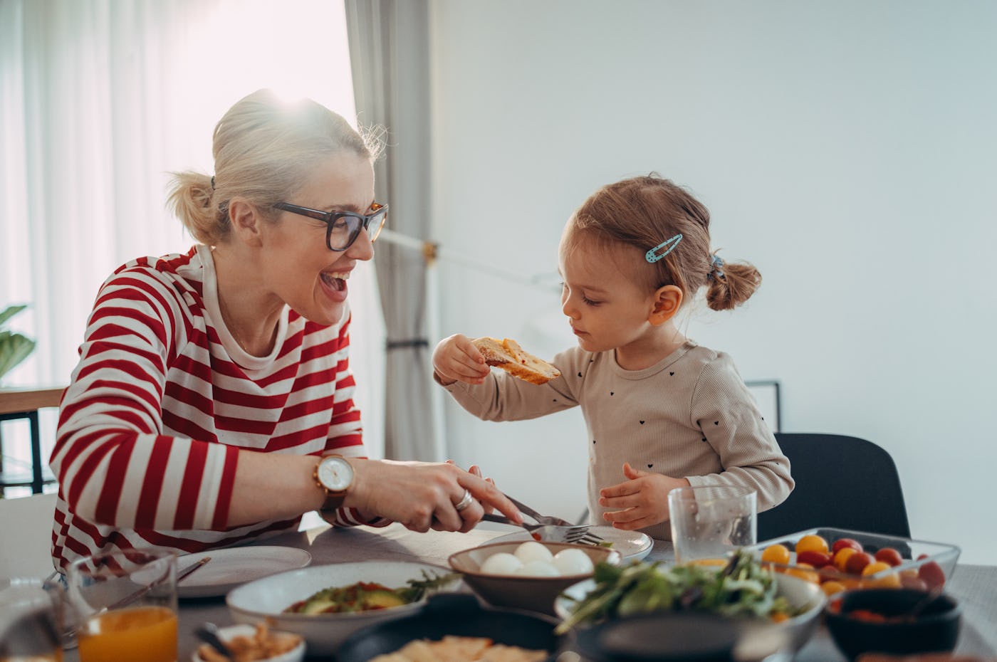 A joyful mother and her her young daughter share a meal together, laughing and interacting at a table filled with delicious food.