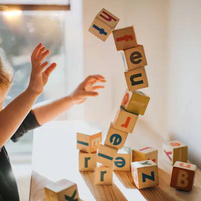 A child plays with colorful wooden blocks, enthusiastically tossing them in the air. Sunlight filters through a window, creating a warm atmosphere.