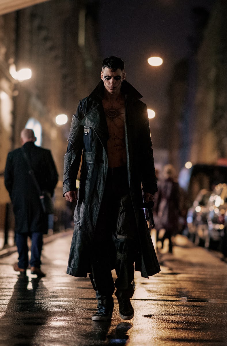 A shirtless man in a long dark coat walks purposefully down a rainy street, with blurred pedestrians...