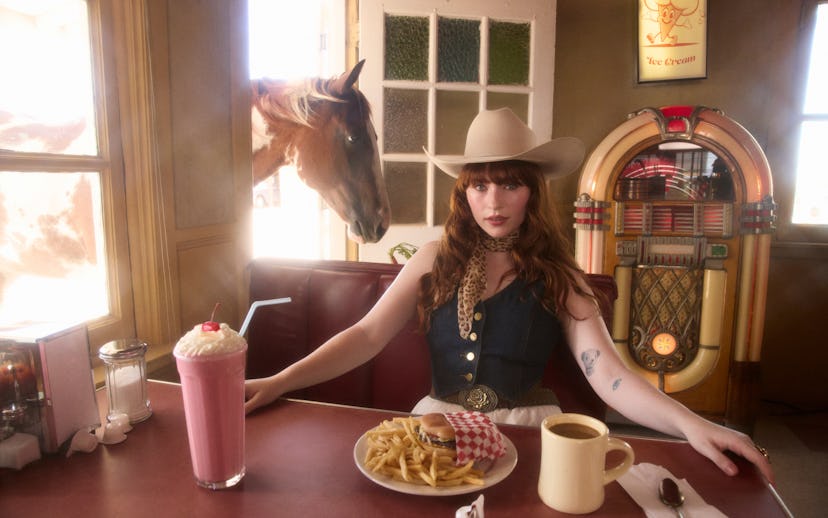 A woman in a cowboy hat sits at a diner table with a milkshake, coffee, and fries, while a horse pee...