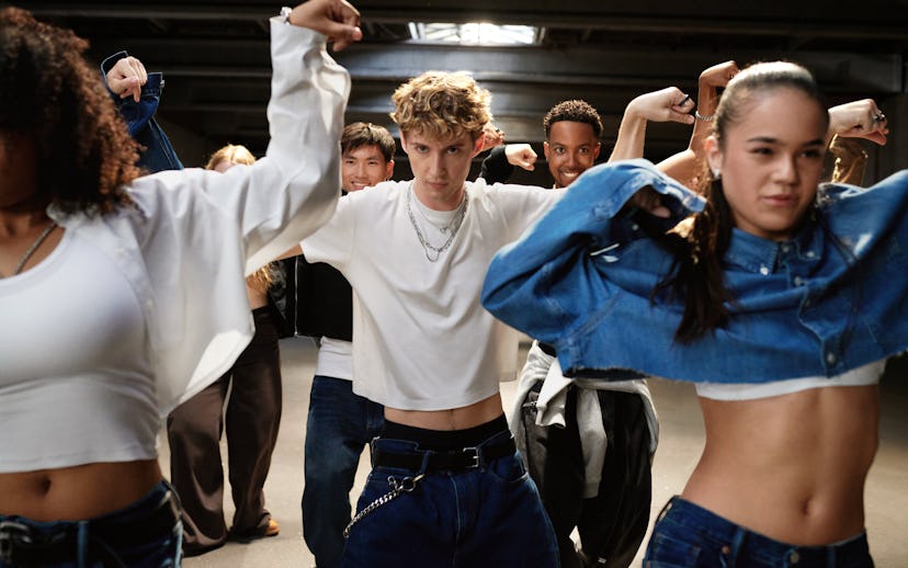 A diverse group of young dancers poses confidently in a dimly lit space, showcasing stylish streetwe...