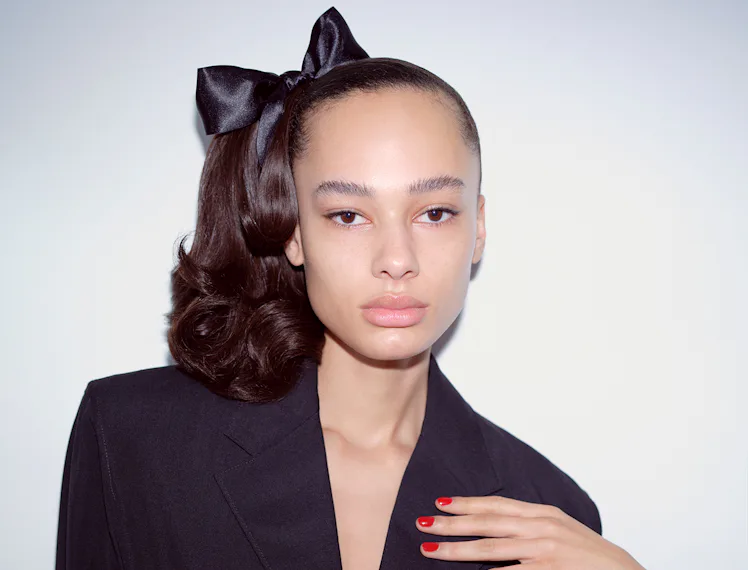 A model with red nails, a fresh face, and brushed brows sports a black silk bow in her sleek and bou...