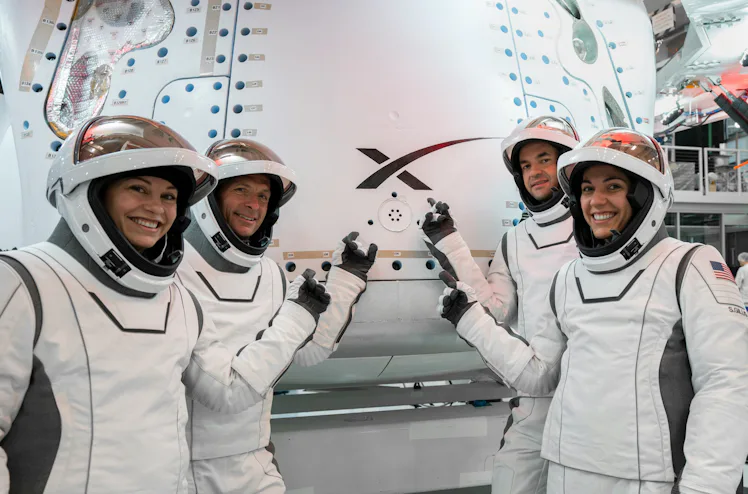 photo of 4 astronauts in white spacesuits, with their visors open, standing in front of a white caps...