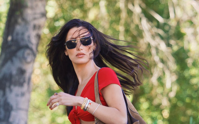 A woman with long hair walks outdoors, wearing a red crop top and denim shorts, accessorized with su...