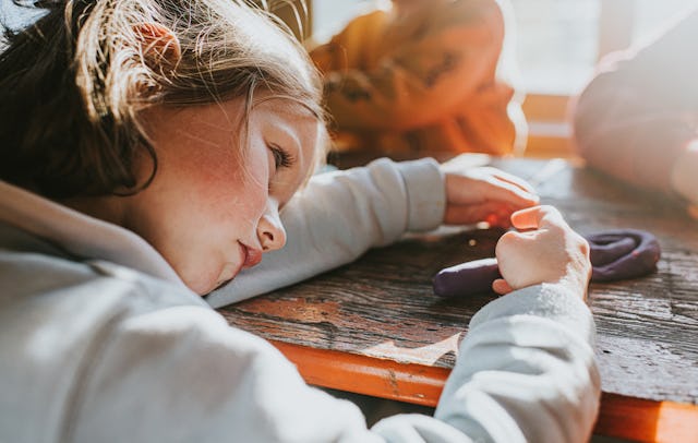 A child rests their head on a wooden table, playing with purple modeling clay. Soft sunlight filters...