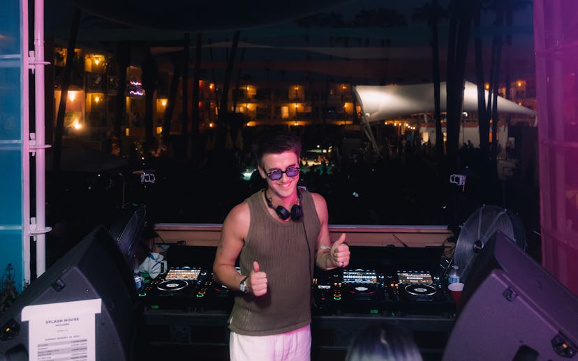 A DJ stands at a booth with thumbs up, wearing sunglasses, in a vibrant outdoor setting with colorfu...