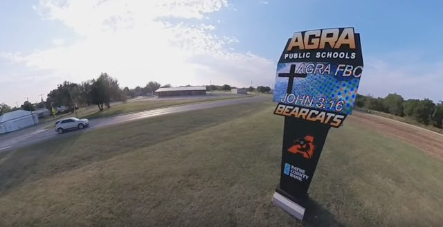 A roadside sign for Agra Public Schools displays "Agra FB," "John 3:16," and "Bearcats," with a car ...