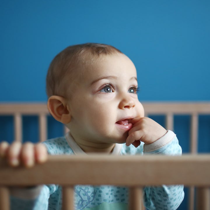 A curious baby with big eyes and a thoughtful expression stands in a crib, holding onto the wooden r...