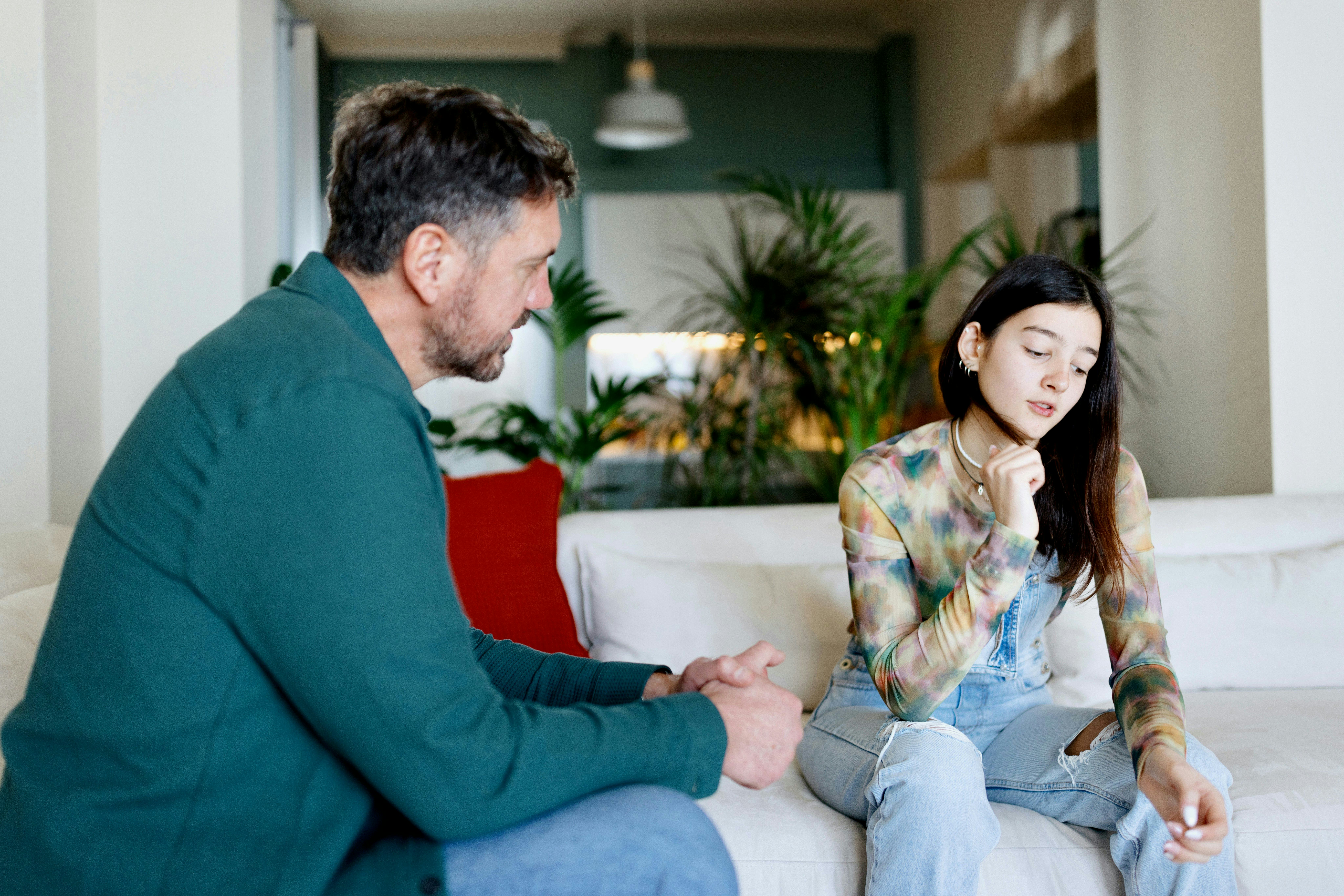 A man and a young woman sit on a sofa in a bright room with plants. The man appears attentive while the woman looks thoughtful, lost in contemplation.