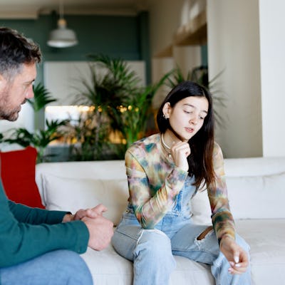 A man and a young woman sit on a sofa in a bright room with plants. The man appears attentive while the woman looks thoughtful, lost in contemplation.