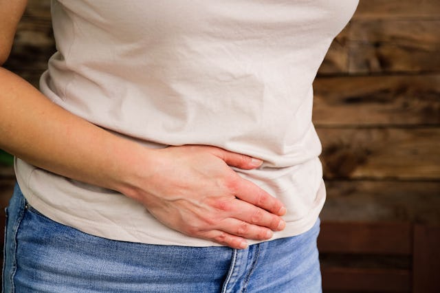 A woman in a light-colored shirt and blue jeans is gently holding her stomach with one hand, express...
