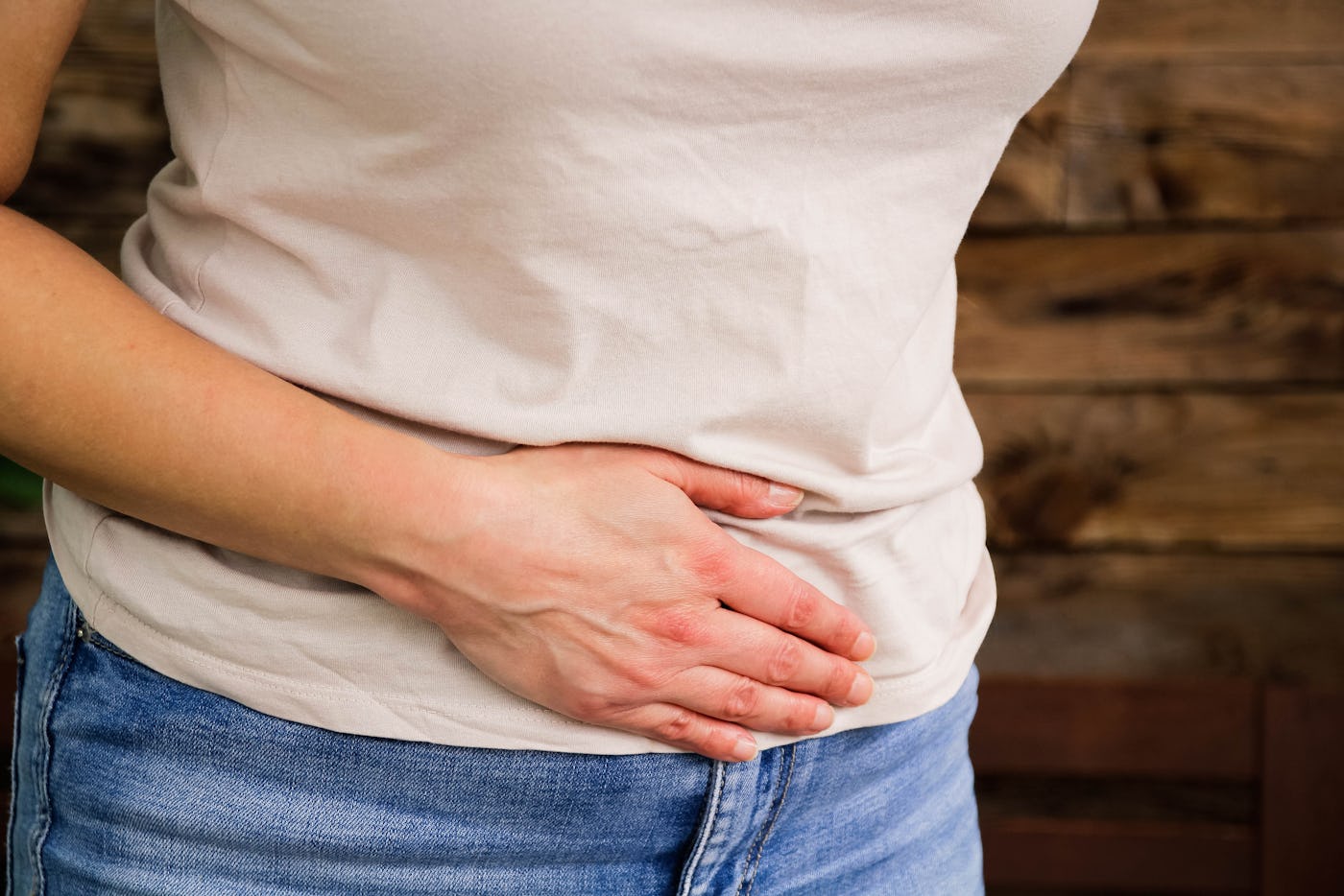 A woman in a light-colored shirt and blue jeans is gently holding her stomach with one hand, expressing discomfort. A wooden background adds warmth to the scene.