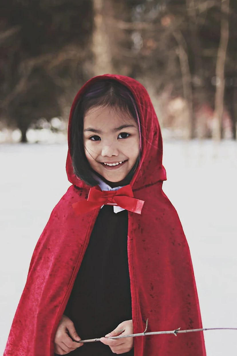 A young girl wearing a vibrant red cape with a bow stands in a snowy landscape, smiling warmly at th...