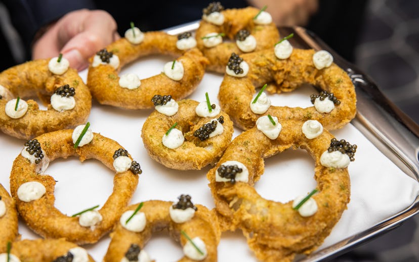 A server presenting a tray of fried onion rings topped with dollops of cream and caviar, garnished w...