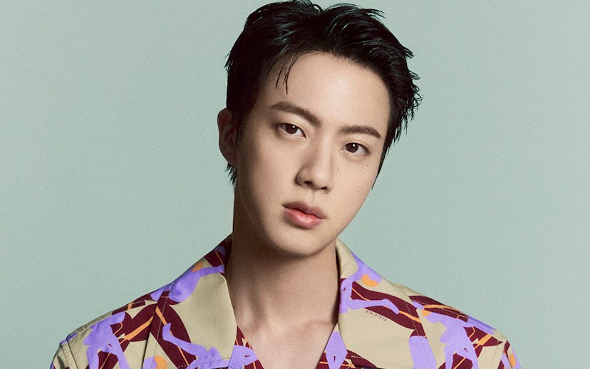 A young man with dark, styled hair wears a colorful short-sleeve shirt with a unique pattern, posing...