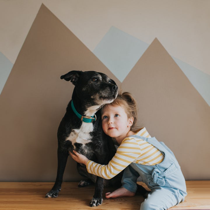 A young girl hugging a black dog against a geometrically patterned wall. Both look off to the side.