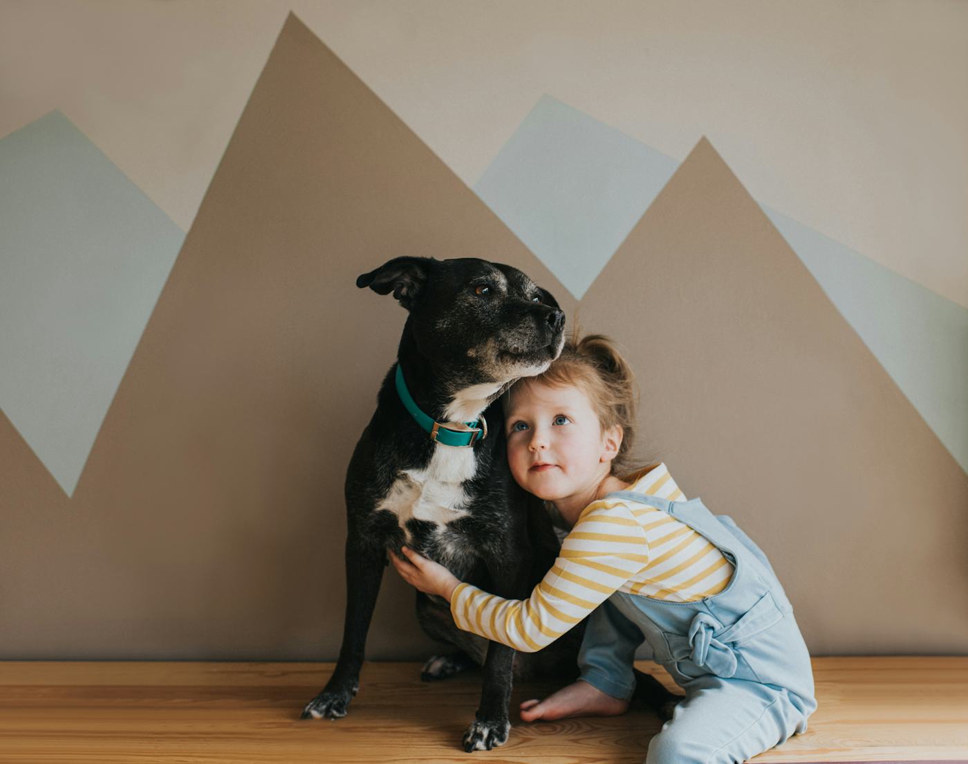A young girl hugging a black dog against a geometrically patterned wall. Both look off to the side.