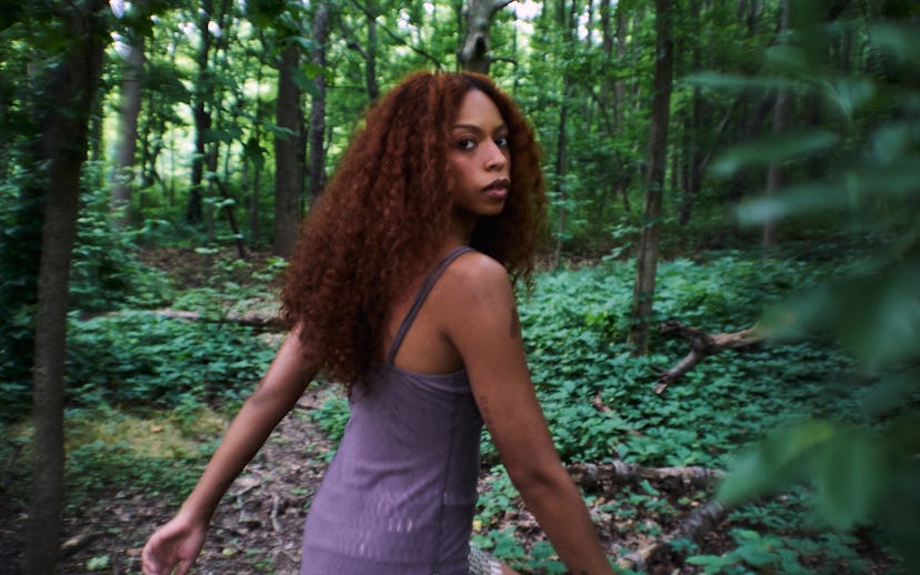 A woman with long, curly hair walks through a lush green forest, turning her head slightly back towa...