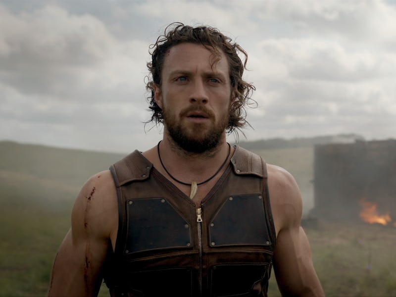 Man with wavy hair and body armor stands in front of a smoldering, burnt structure in a grassy field...
