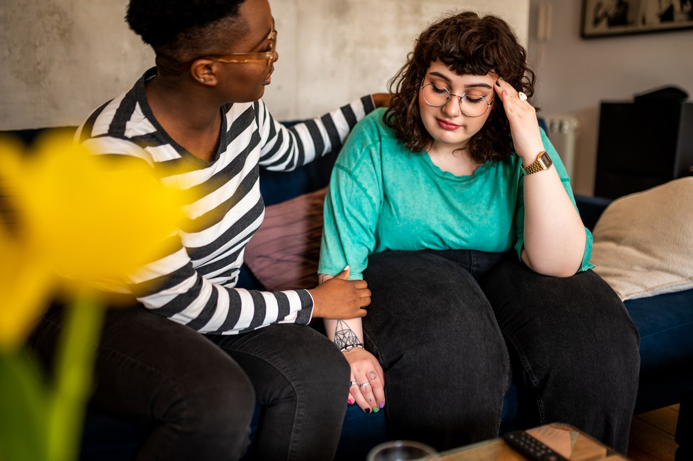 Two women sit together on a couch in a cozy living room. One comforts the other, who appears to be distressed, highlighting emotional support.