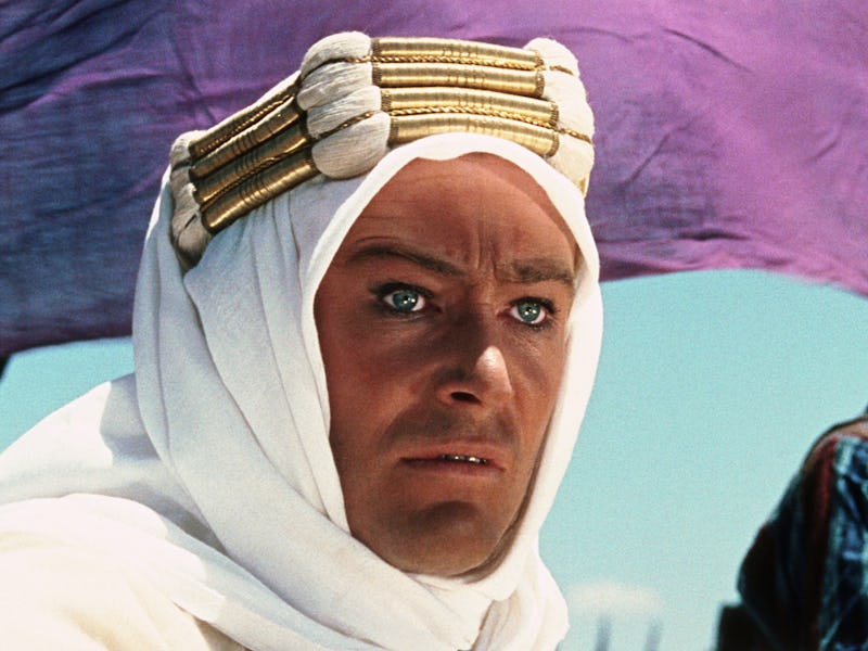 A close-up of a man wearing a white robe and headdress, with a serious expression, set against a bri...