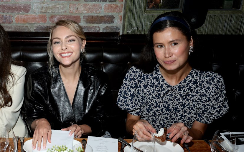Two women sit at a dining table, smiling. One wears a black leather jacket, while the other sports a...