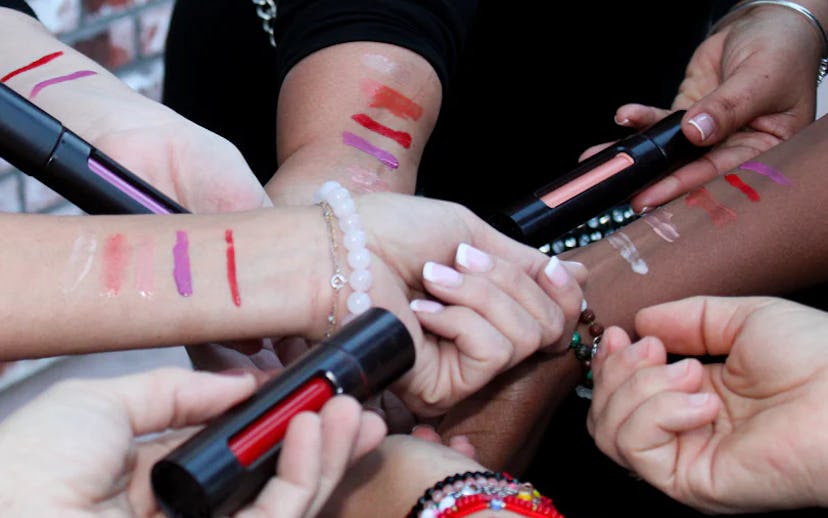 A group of hands displaying various lip products, with colorful swatches on their arms against a bri...