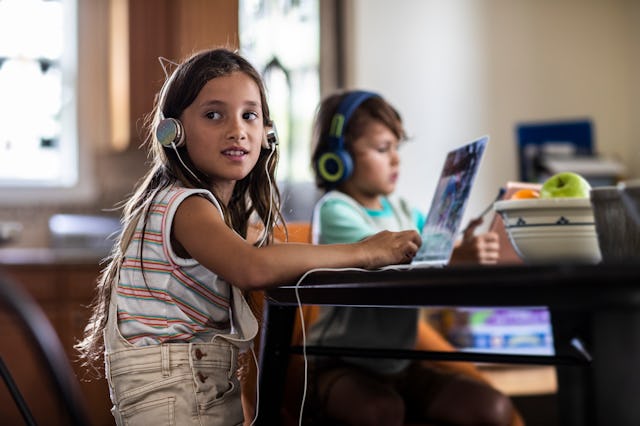 A young girl with long hair and headphones smiles while using a laptop at a table. A boy with headph...