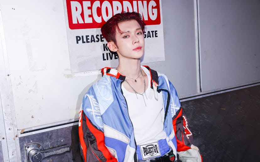 A person with striking red hair poses confidently in a colorful jacket, standing against a sign that...