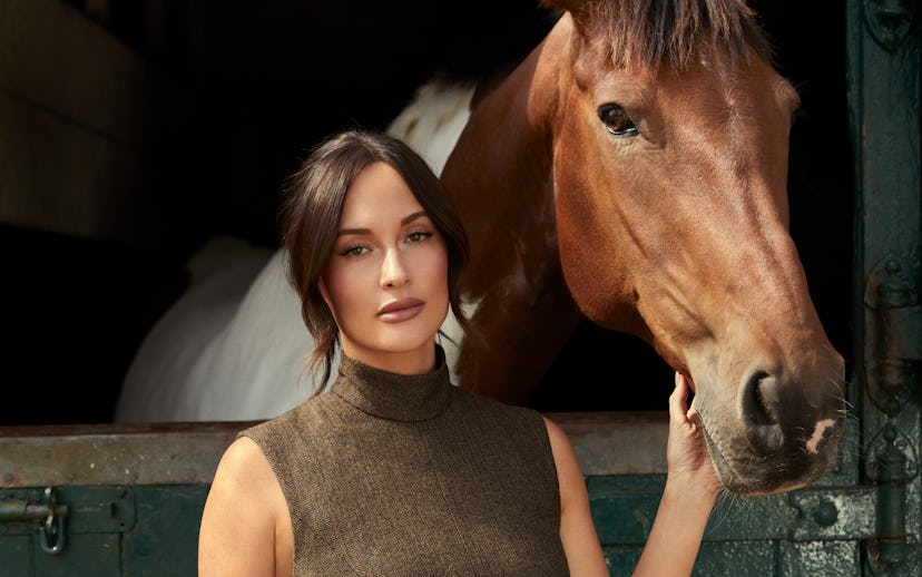 A woman in a fitted brown dress stands beside a brown horse at a stable, with a green wooden door in...