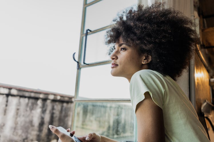 Young woman looking out window holding her phone after reading her October 2024 horoscope.