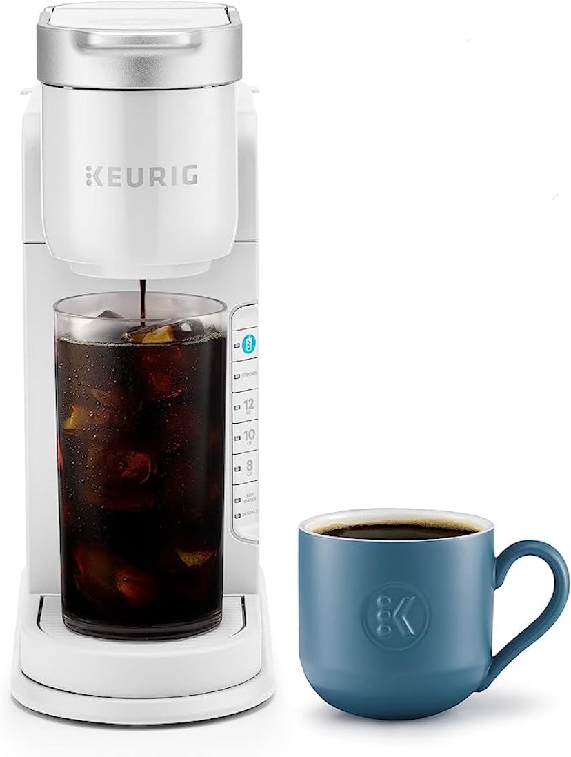 A white Keurig coffee maker brews iced coffee into a glass container, accompanied by a blue ceramic ...