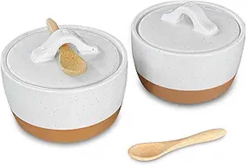 Two ceramic storage bowls with white speckled lids and brown bases, accompanied by a small wooden sp...