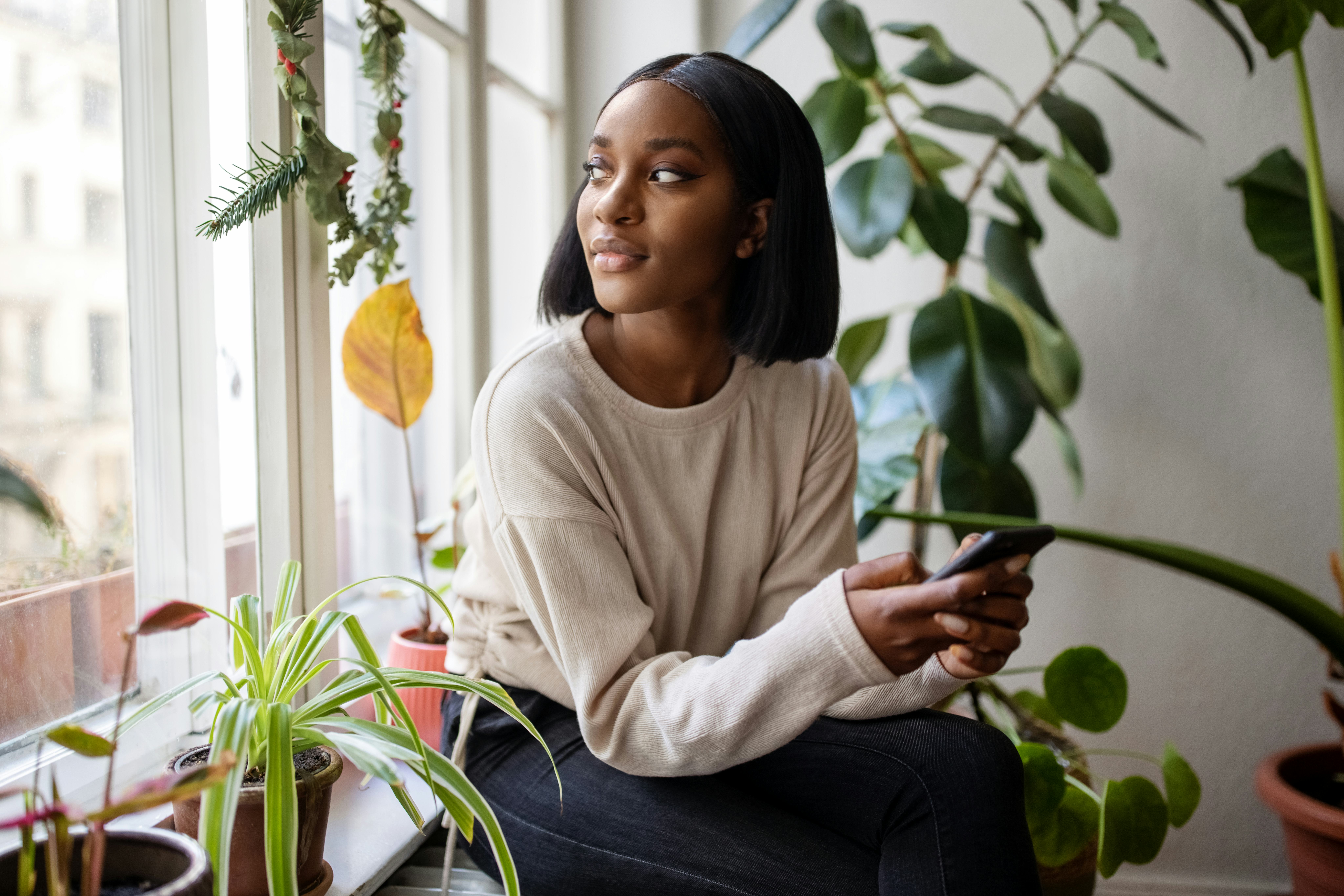 Young woman staring out window among plants after reading her October 2024 horoscope.
