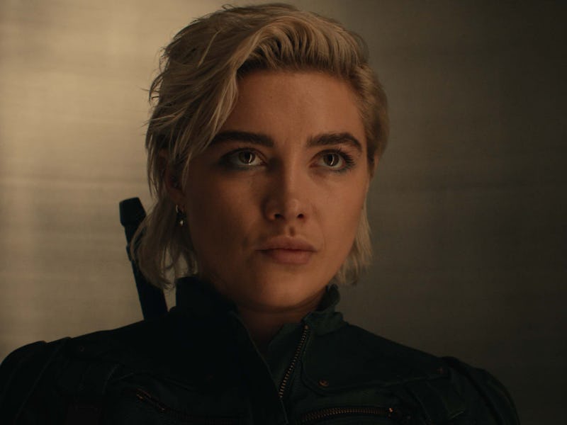 A close-up of a young woman with short blonde hair and a serious expression, wearing a dark outfit, ...
