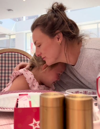 A woman gently embraces a young girl, both showing emotion, while seated at a table adorned with plates and condiments in a bright, airy setting.
